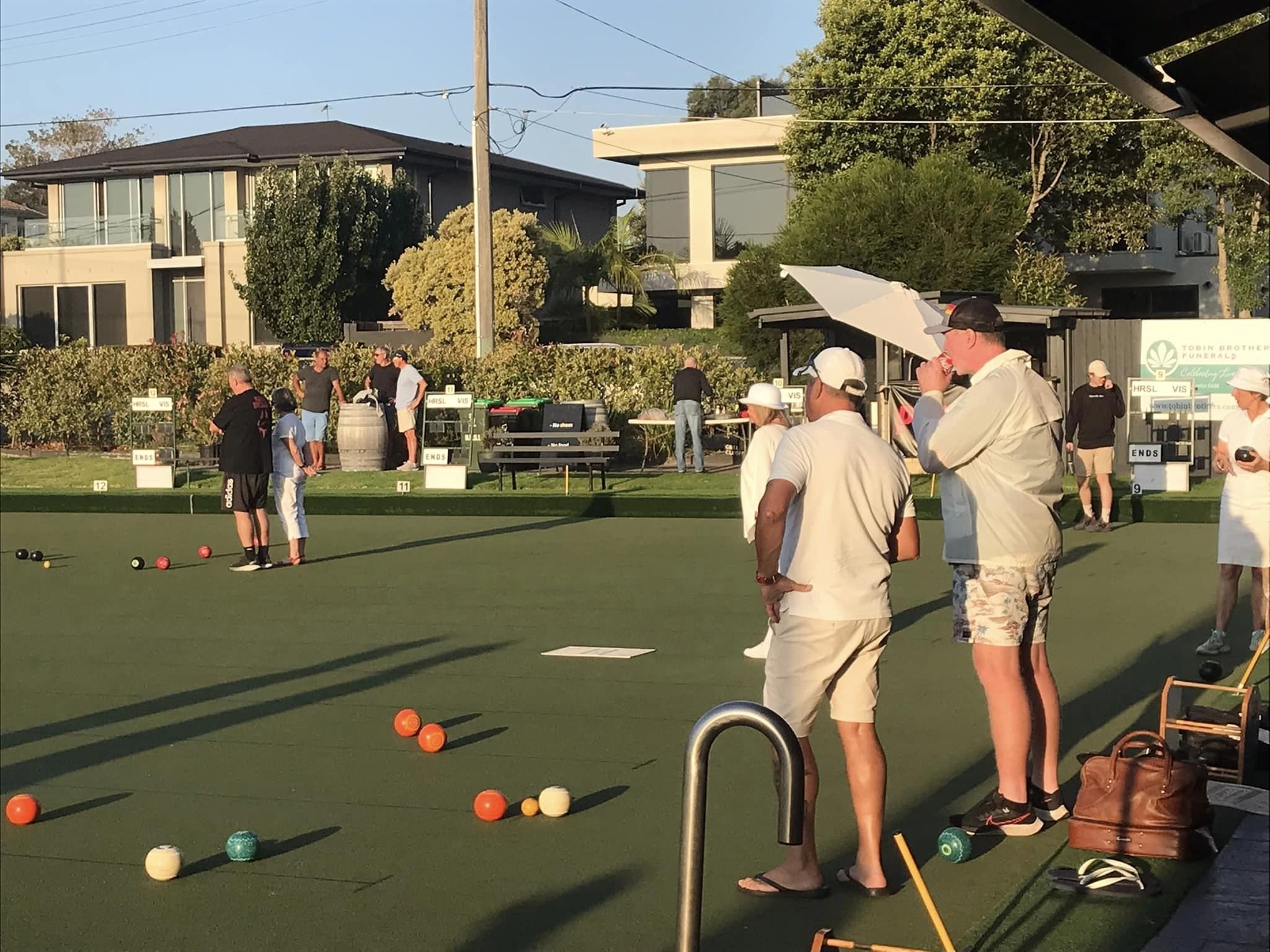 Bowls spread across the green on a sunny evening while players chat by the bank
