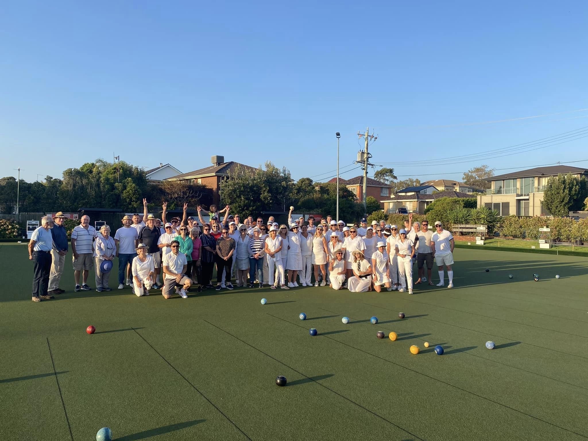 Members and guests in whites on the green at a barefoot bowls night, bowls scattered across the rink