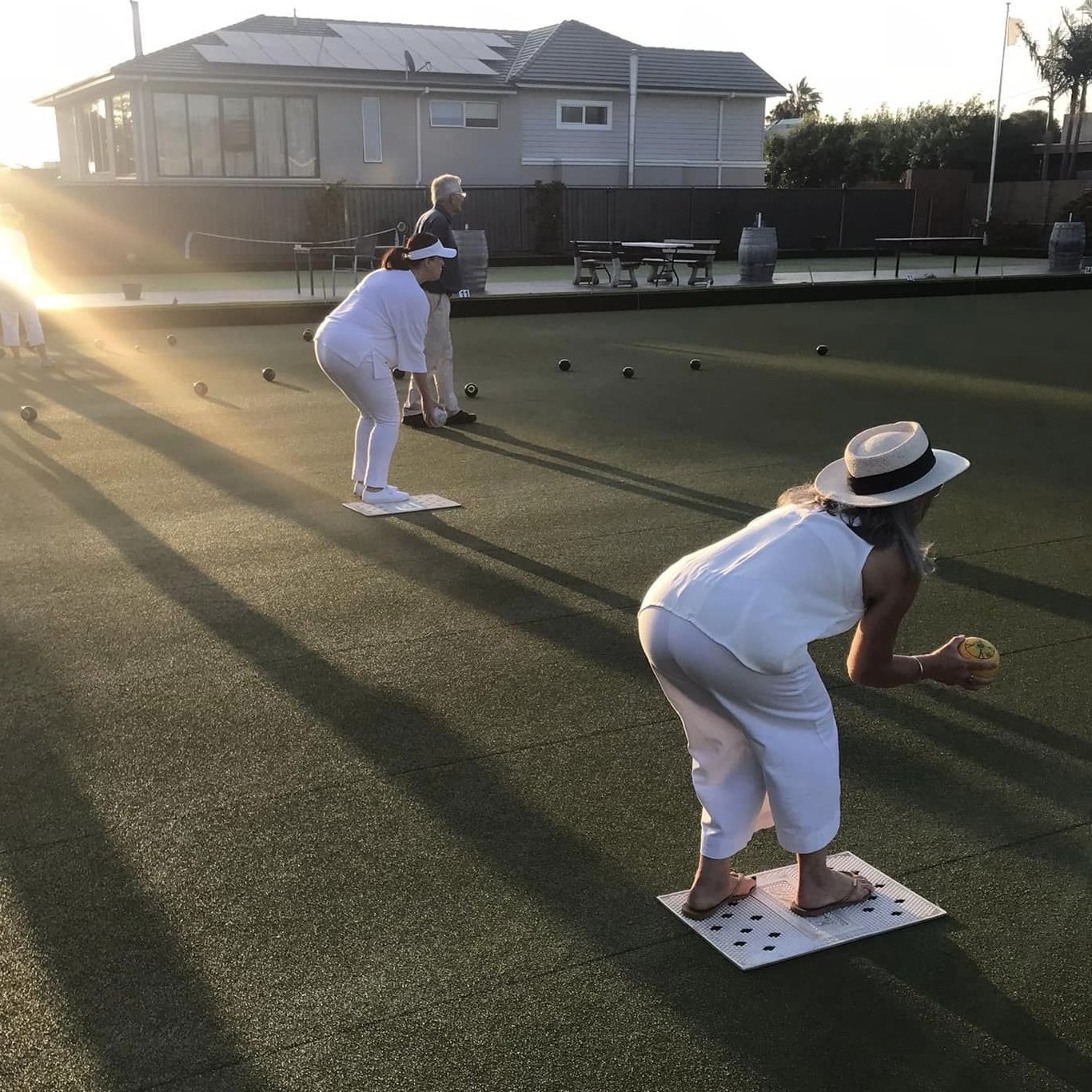 A bowler in whites delivering on the mat as the sun drops behind the clubhouse